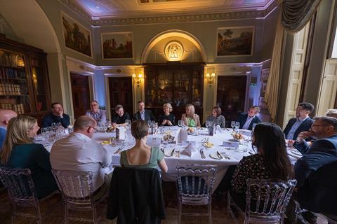 Group photo of 16 attendees sitting around the event's roundtable. They sit in a low-lit room, surrounded by bookcases and paintings on the walls, with open-shuttered windows letting in light from the right.